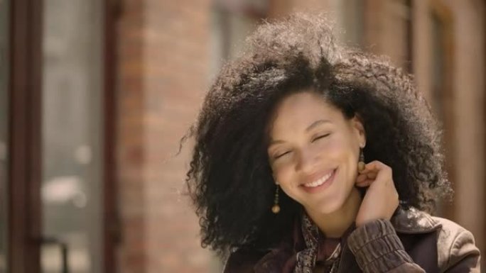 年轻的非洲裔美国妇女看着相机微笑的肖像。穿着棕色皮夹克的黑发女人在模糊的建筑背景下摆姿势。特写。慢动
