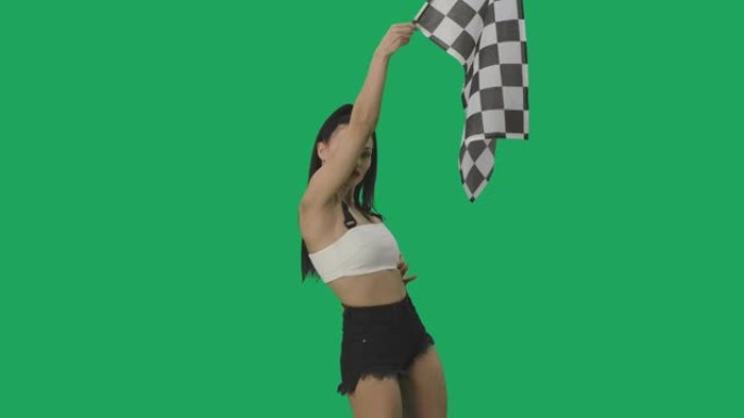 穿着短裤的性感黑发女郎挥舞着黑白方格赛旗，标志着比赛的开始。年轻女子在绿屏背景下摆姿势特写。慢动作就