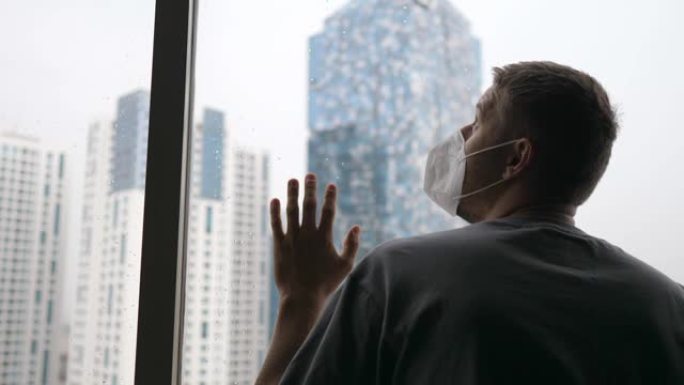 一个戴着面具的成年白人男子透过摩天大楼的窗户看。新型冠状病毒肺炎大流行引起的抑郁症