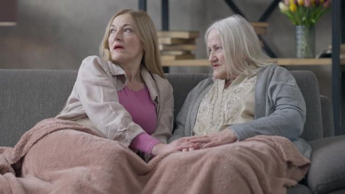 两个白人高级妇女坐在沙发上手牵手交谈。严重的女性退休人员在室内互相支持的问题的中等镜头肖像。友谊与衰