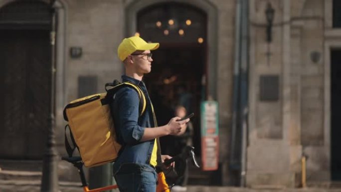 一个年轻快乐的送货员正拿着智能手机去。他带着一辆自行车。他戴着黄色的帽子和眼镜。他肩上背着一个黄色的