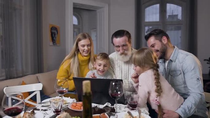 快乐的家庭在电脑上看着美丽的照片，这些照片显示了他们尊敬的爷爷，一起坐在桌子旁