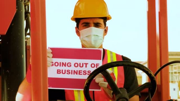 年轻的男性建筑工人看着戴着防护面罩的摄像头，拿着一辆大尺寸叉车的方向盘，举着 “退出营业” 的牌子
