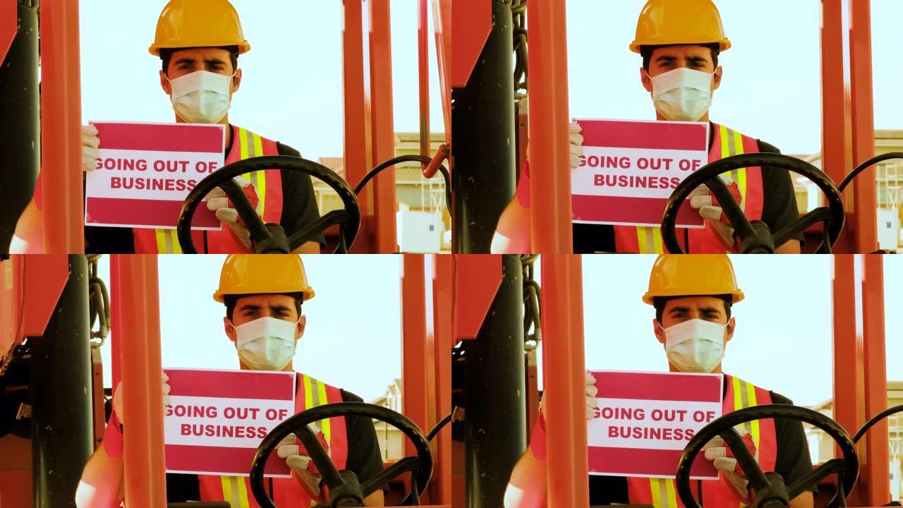 年轻的男性建筑工人看着戴着防护面罩的摄像头，拿着一辆大尺寸叉车的方向盘，举着 “退出营业” 的牌子