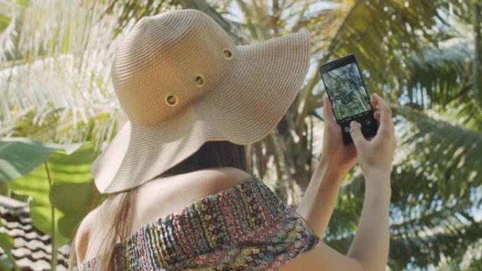 无法辨认的女人在蓝天背景上拍摄绿色棕榈树的背面视图。戴帽子的不露面的女孩制作热带自然慢动作的手机照片