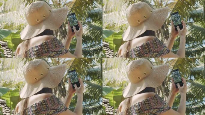 无法辨认的女人在蓝天背景上拍摄绿色棕榈树的背面视图。戴帽子的不露面的女孩制作热带自然慢动作的手机照片