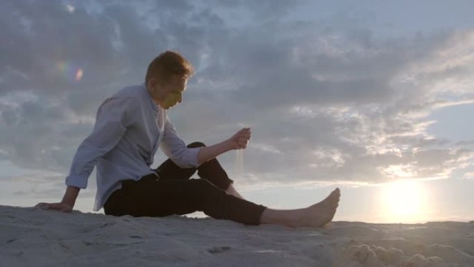 英俊的悲伤的家伙在日落天空的背景下坐在沙滩上，在手指间倒沙。概念男模。情绪。慢动作