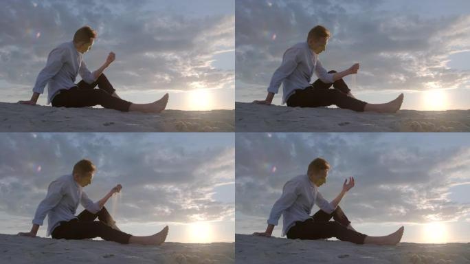 英俊的悲伤的家伙在日落天空的背景下坐在沙滩上，在手指间倒沙。概念男模。情绪。慢动作