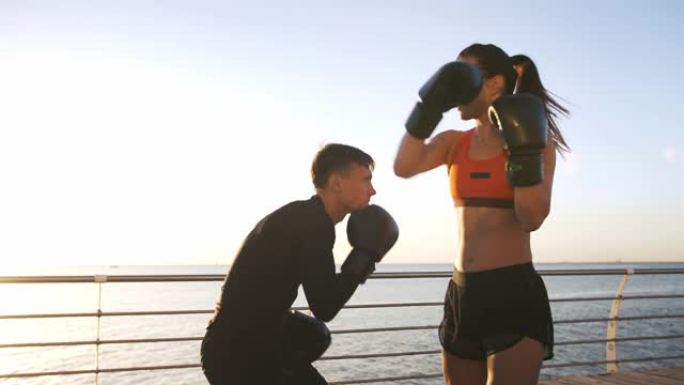 穿着运动服的年轻女性正在和她熟练的教练一起拳击。他们在海上海滨长廊训练时躲避拳头