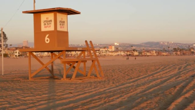 标志性的复古木制橙色救生员守望塔在沙质加利福尼亚太平洋海滩上被日落光线照亮。私人度假屋和地平线上的山