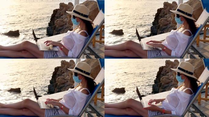 年轻女子戴着防护面具，戴着太阳帽和夏装，在笔记本电脑上工作，坐在海边的日光浴躺椅上。远程工作。新常态