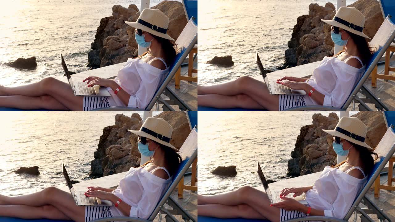 年轻女子戴着防护面具，戴着太阳帽和夏装，在笔记本电脑上工作，坐在海边的日光浴躺椅上。远程工作。新常态