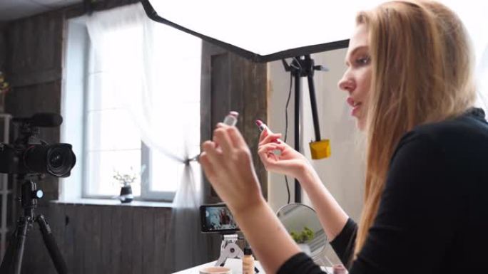 Vlogger女性在嘴唇上涂抹口红。美容博主女人在三脚架上的相机上拍摄日常化妆常规教程。有影响力的金