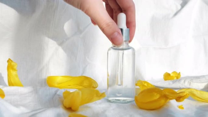 透明玻璃瓶化妆油的特写镜头，白色背景上被黄色郁金香花瓣包围。概念。美容美容，女人手拿走小瓶