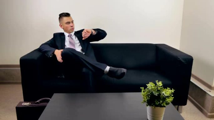 一个穿着西装的年轻商人正坐在沙发上等待采访，看着他的手表。这个人想找份工作。