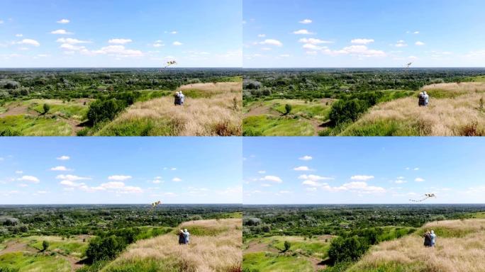 航空。幸福的年轻夫妇在绿色山谷和蓝天的美丽风景的背景下，放飞五颜六色的风筝，玩得开心。后视图。夏季晴