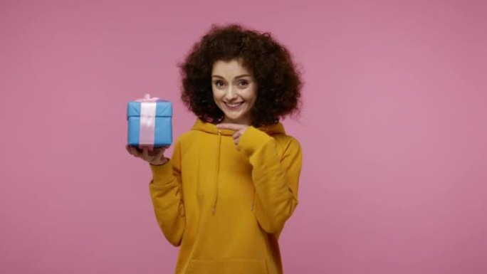 这礼物给你!迷人的快乐女孩非洲发型，连帽衫指着包装好的礼品盒，微笑着