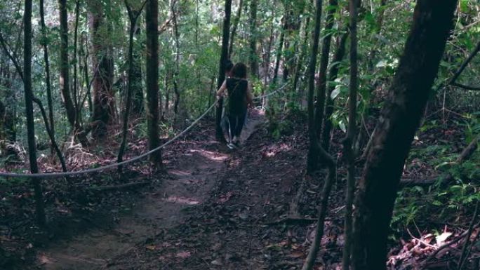 徒步旅行两个女孩背着背包在雨林丛林中行走，从后视图抓住人行道上的强力绳索