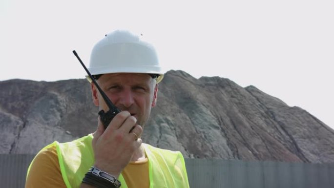 年轻的建筑工人穿着夹克和安全帽，在沙山的背景下用对讲机说话