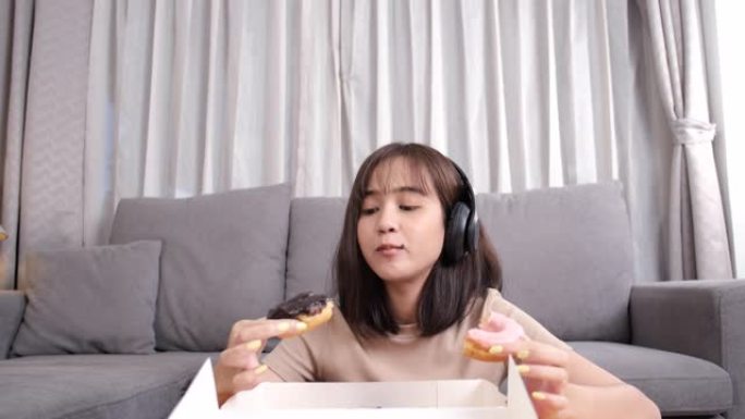 年轻的亚洲女性外卖在沙发上吃甜甜圈垃圾食品，在客厅听音乐，快餐送货服务安全，并通过社交距离概念阻止冠