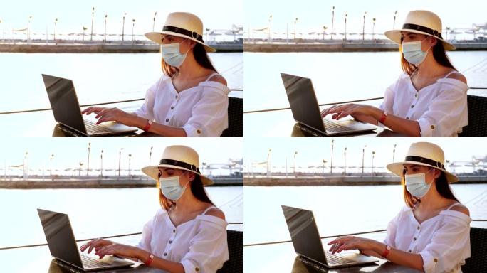 年轻女子戴着防护面具，戴着遮阳帽和夏装，在笔记本电脑上工作，坐在海边的空堤上。远程工作。新型冠状病毒