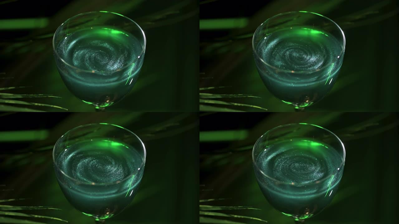 深绿色背景上带有绿色鸡尾酒的酒杯，带有植物叶子。库存镜头。带有闪亮颗粒的绿色酒精饮料的特写