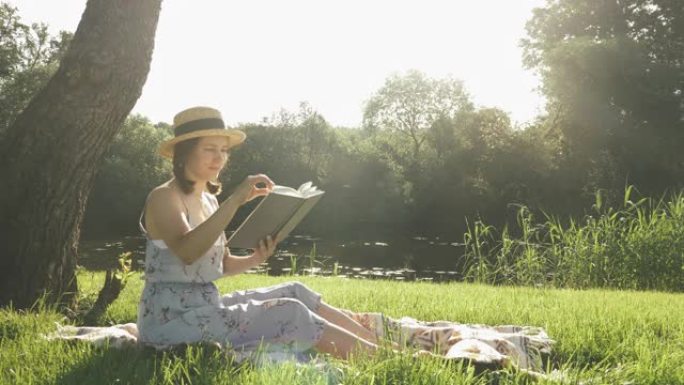 微笑的快乐女孩戴着帽子，穿着看书日记坐在河边大树下的草地上。享受周末假期的女人。可爱的女士坐在公园的