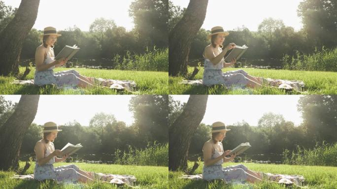 微笑的快乐女孩戴着帽子，穿着看书日记坐在河边大树下的草地上。享受周末假期的女人。可爱的女士坐在公园的
