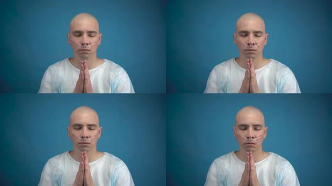秃头的年轻人和肿瘤学人看着相机，在蓝色背景上祈祷。病人双手合十祈祷。化疗导致脱发。