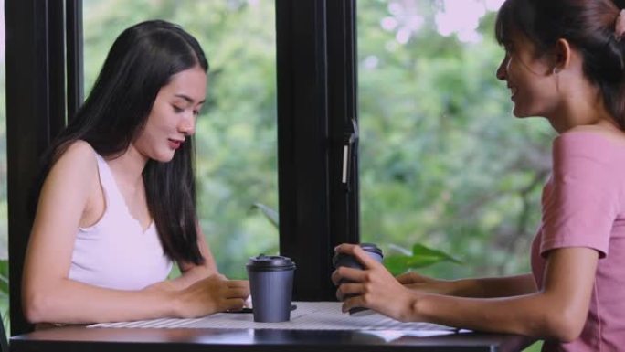 亚洲女人和一个朋友在咖啡店里玩电话，喝热咖啡。