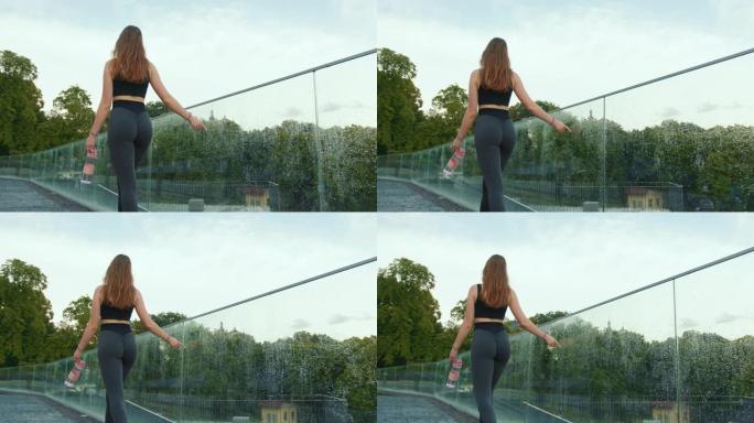一个运动的女孩正在沿着玻璃栏杆走路和奔跑。她穿着紧身裤，手里拿着一个水瓶。从后面射击。4K