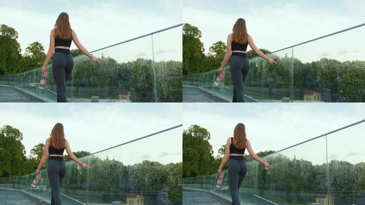 一个运动的女孩正在沿着玻璃栏杆走路和奔跑。她穿着紧身裤，手里拿着一个水瓶。从后面射击。4K