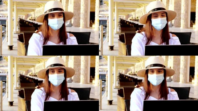 年轻女子戴着防护面具，戴着遮阳帽和夏装，在笔记本电脑上工作，坐在海边的空堤上。远程工作。新型冠状病毒