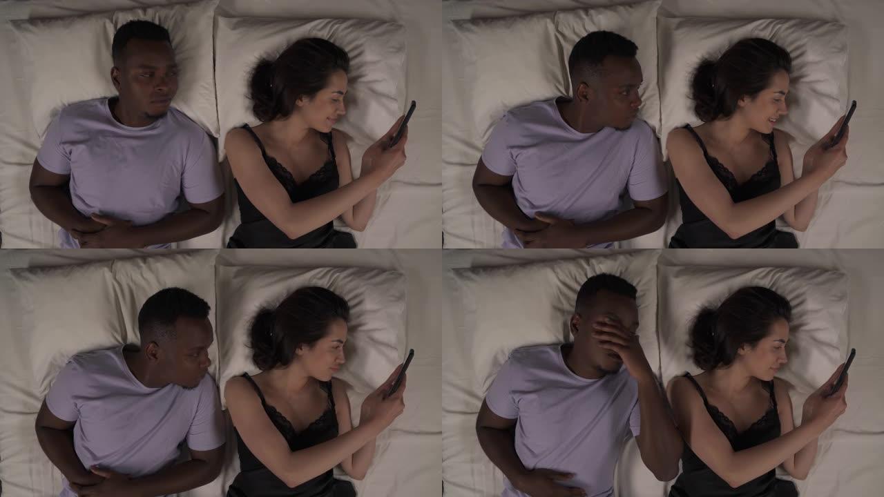 混血夫妇晚上躺在床上的俯视图。白人女孩盯着手机，微笑着。黑人男子偷看屏幕，做出面部表情手势