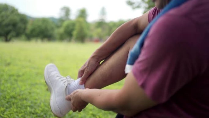 亚洲男子在公园慢跑运动时脚踝受伤坐在树干绿草的背景上，脚踝疼痛，关节韧带问题，意外事故，腿部肌肉肌腱