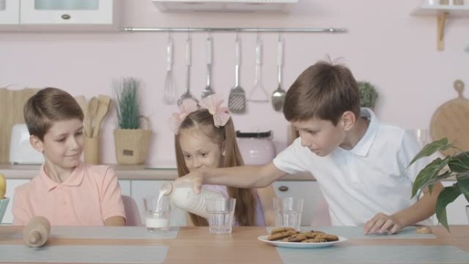 小绅士在厨房里把牛奶倒进玻璃杯的画像。三个孩子坐在家里的餐桌旁吃午饭。双胞胎男孩和女孩在室内一起休息