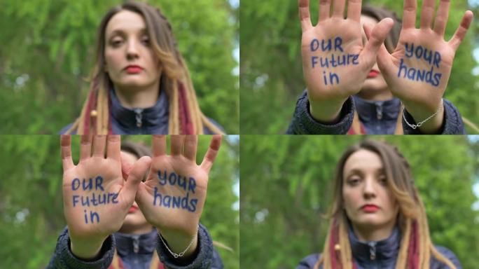 梳着长发的现代严肃女孩在绿树背景上举起手，上面写着“我们的未来在你手中”的口号。责任，气候变化概念