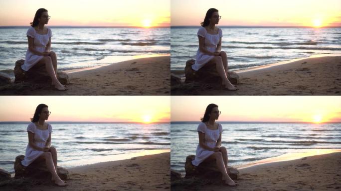 一名年轻女子坐在海边的一块石头上，耳朵里戴着耳机。日落时一个穿着白色连衣裙的女孩戴着耳机听音乐。