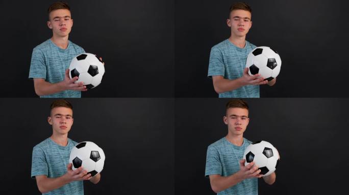 少年男孩的肖像站在足球上，双手握着，在深灰色背景的镜头前，面带微笑。足球迷，球员。青少年，运动。生活