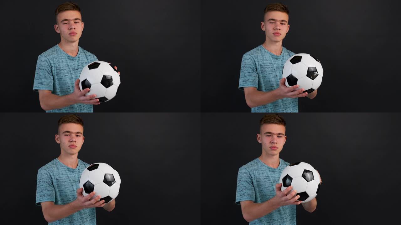 少年男孩的肖像站在足球上，双手握着，在深灰色背景的镜头前，面带微笑。足球迷，球员。青少年，运动。生活