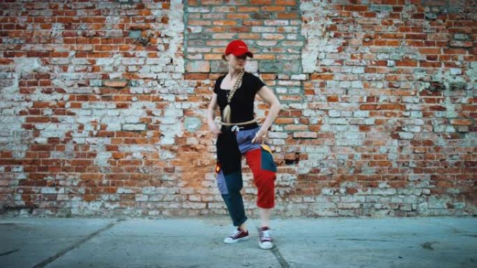 开朗的年轻女子戴着帽子，在城市砖墙旁边的街道上积极跳舞。穿着五颜六色的裤子。127bpm股票视频