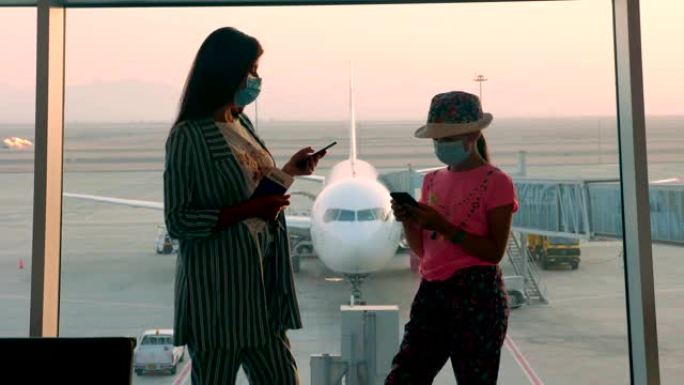 戴着面具，戴着护照和机票的女人和少女在机场的全景窗前使用手机，在跑道和大飞机视野。冠状病毒疫情结束后