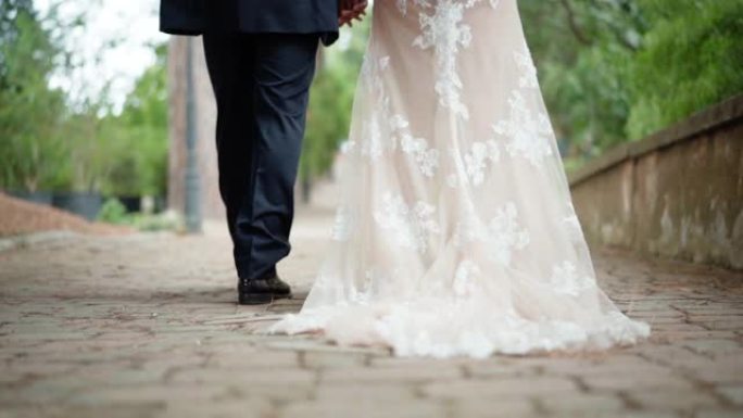 沿着公园古老的鹅卵石延伸的牛奶色时尚蕾丝婚纱的特写镜头。结婚典礼结束后，刚结婚的夫妇在花园里慢慢地牵