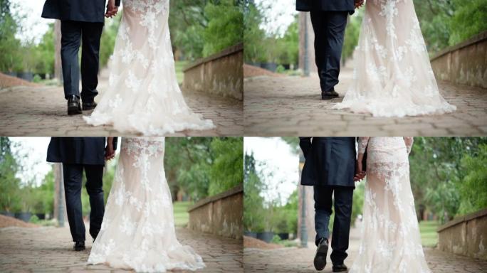 沿着公园古老的鹅卵石延伸的牛奶色时尚蕾丝婚纱的特写镜头。结婚典礼结束后，刚结婚的夫妇在花园里慢慢地牵