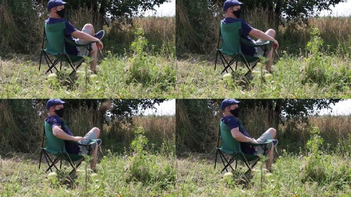 一个蒙面人坐在野营椅上。我出去在城市公园呼吸新鲜空气。在流行和自我隔离期间散步和休息。最后一个计划。