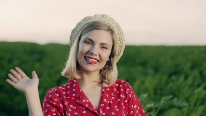 美丽的肖像迷人的金发女人挥手，微笑，问候。绿色自然背景上戴着草帽的女士。愉快的女性外表，善良的微笑。