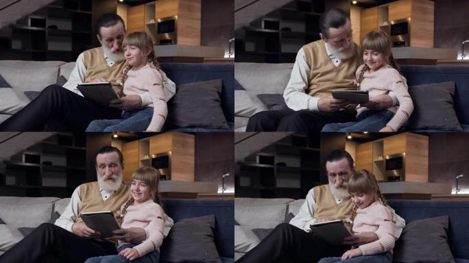 美丽的12岁女孩，有滑稽的辫子，和她尊敬的大胡子祖父坐在沙发上，在i-pad的屏幕上观看