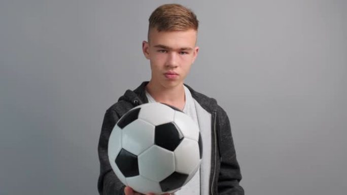 肖像少年男孩认真的旋转不耐烦地将足球从手到手扔出去，并在灰色背景下在电视上观看比赛。青少年和体育。生