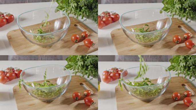 芝麻菜的绿色湿叶落入玻璃碗中。在西红柿，盐和胡椒粉的背景下准备沙拉蔬菜。全景慢动作。全高清视频，24