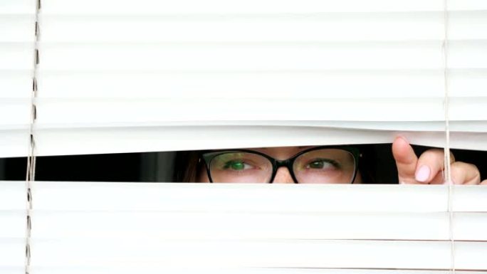戴眼镜的女人透过白色百叶窗看着某人